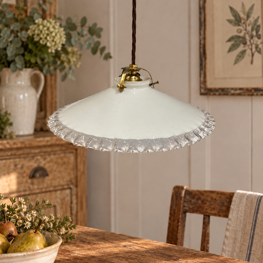 A vintage-style pendant light with a white glass shade and a scalloped clear crystal-like trim, hanging over a wooden dining table in a warm, rustic room.