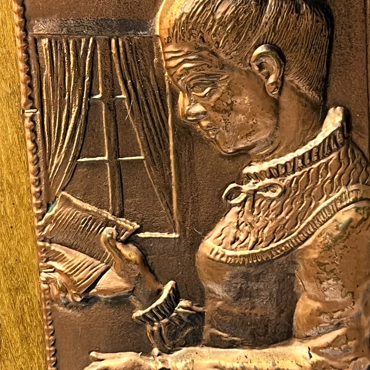 Close-up of a copper relief sculpture depicting a woman combing her hair in front of a window with curtains.