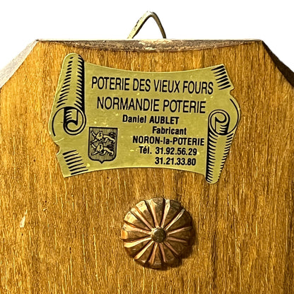 Close-up of a wooden decorative plaque or box front with a brass-colored metal label reading 'POTERIE DES VIEUX FOURS NORMANDIE POTERIE Daniel AUBLET' and a round metallic medallion below.