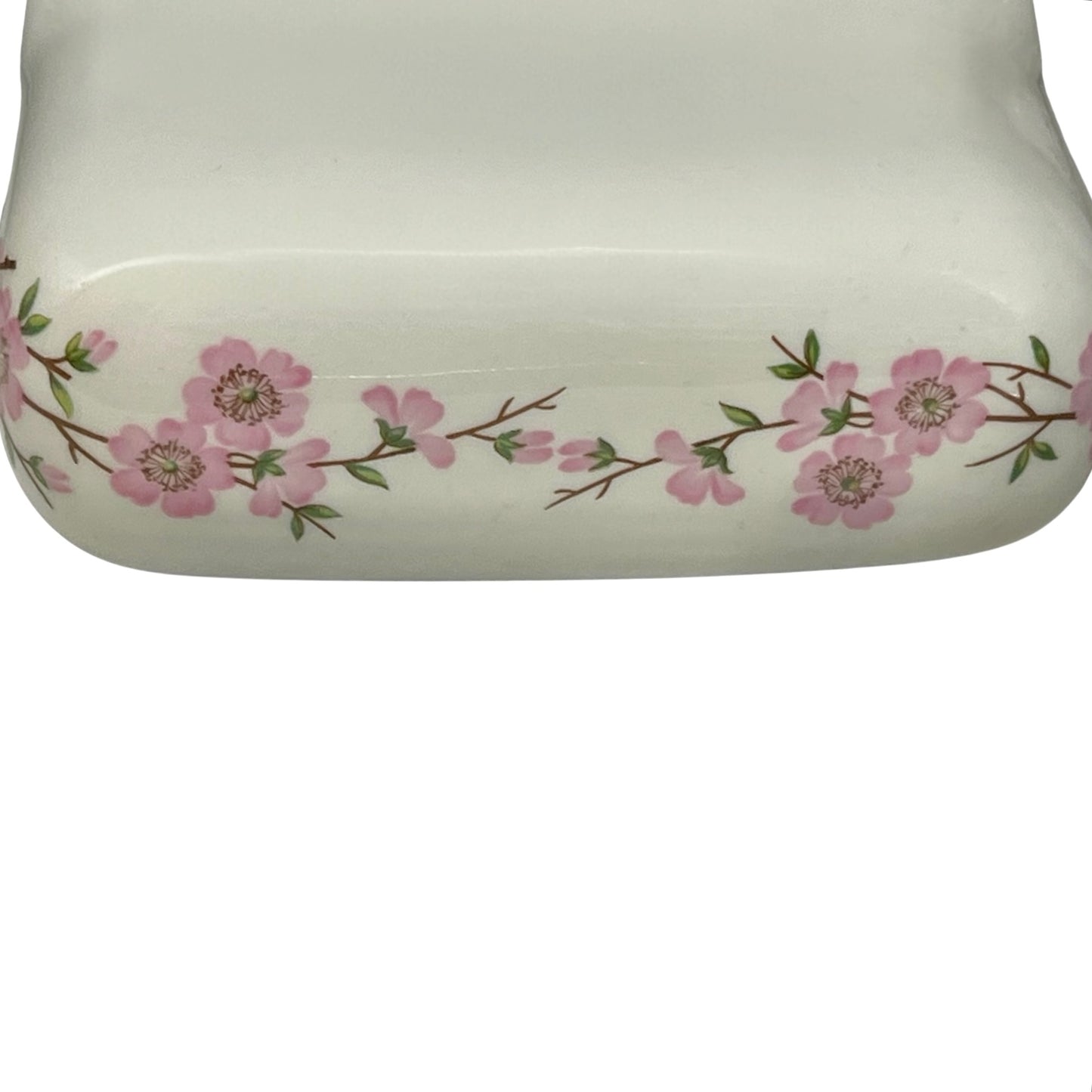 Close-up of a glossy white ceramic container or vase featuring a delicate pink floral branch pattern along the rim, set against a plain white background.