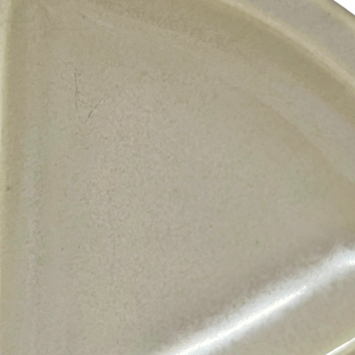 A close-up of a light beige ceramic surface, likely a plate or dish, showing a slightly textured glaze with a bright reflection along the bottom right edge.