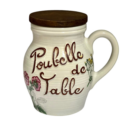 A cream-coloured ceramic table jar with a dark wooden lid, featuring the French text 'Poubelle de Table' in brown script and pink floral decoration on a white background