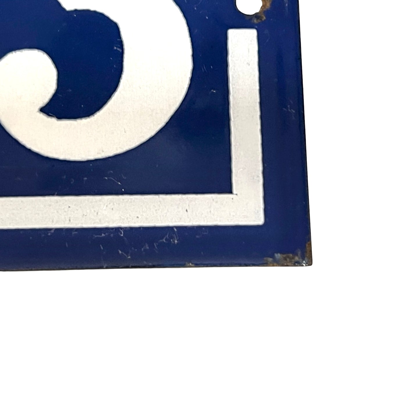 A close-up of a vintage-style blue enamel sign with a white painted letter or symbol, showing some wear, rust, and a small hole near the top edge against a white background.