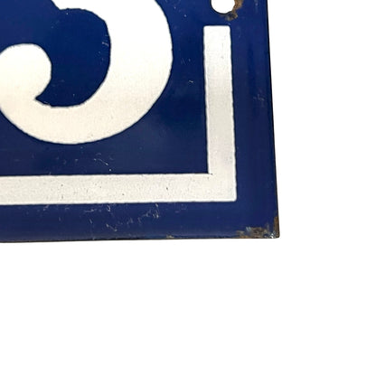 A close-up of a vintage-style blue enamel sign with a white painted letter or symbol, showing some wear, rust, and a small hole near the top edge against a white background.