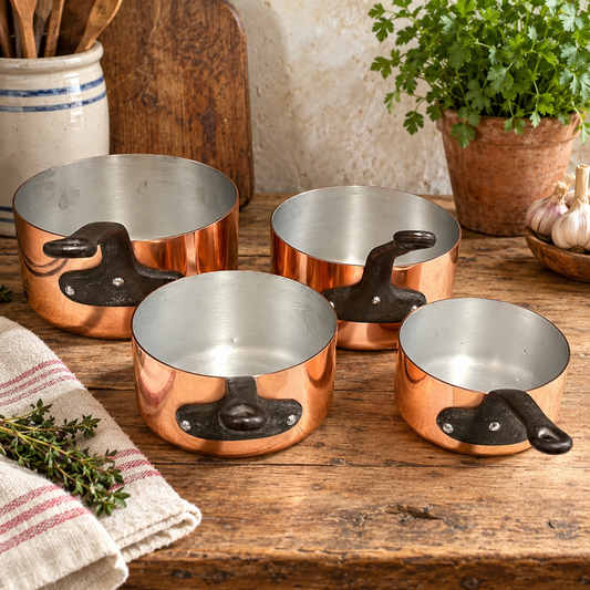 Four copper saucepans with black handles arranged on a rustic wooden table, with kitchen props including herbs, garlic, a potted plant, and a striped cloth nearby.
