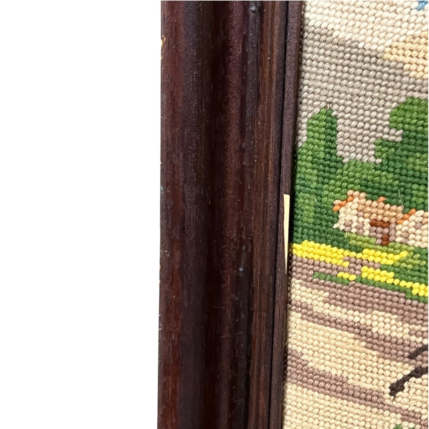 Close-up of a framed needlepoint canvas showing a dark brown wooden frame on the left and part of a stitched landscape design with green trees, yellow flowers, and a beige path.