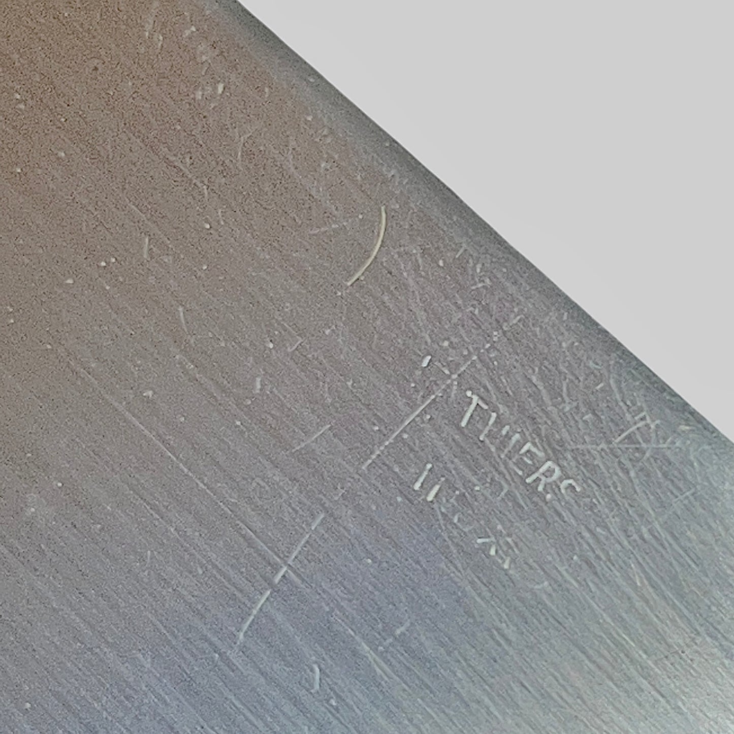 A close-up of a brushed metal surface with visible scratches and faint stamped text, shown against a light gray background.