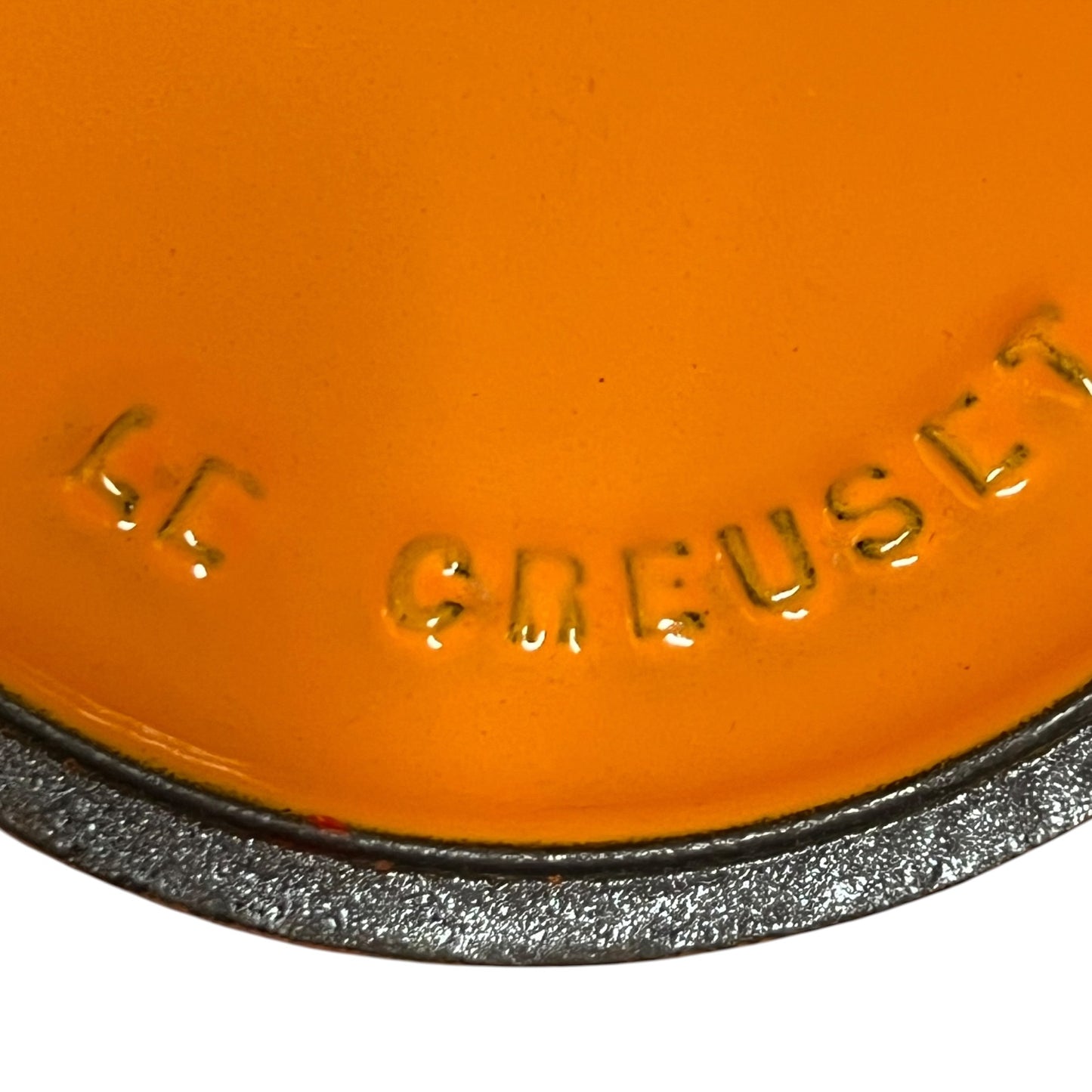Close-up of an orange enamel item with embossed 'LE CREUSET' lettering and a dark gray cast-iron rim, on a white background.