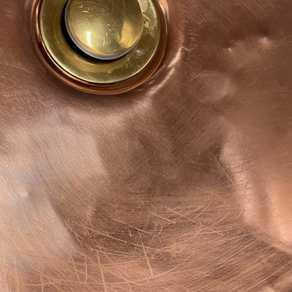 Close-up of a copper sink basin with a brass drain and pop-up cover, showing the brushed metal surface and scratches.