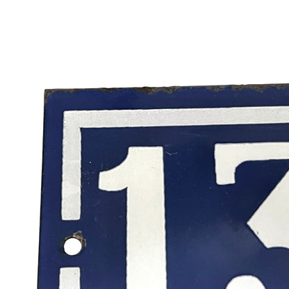 Close-up of a blue and white enamel-style sign showing part of the number 133 with a small round hole near the lower left and a worn, rusted edge along the top.