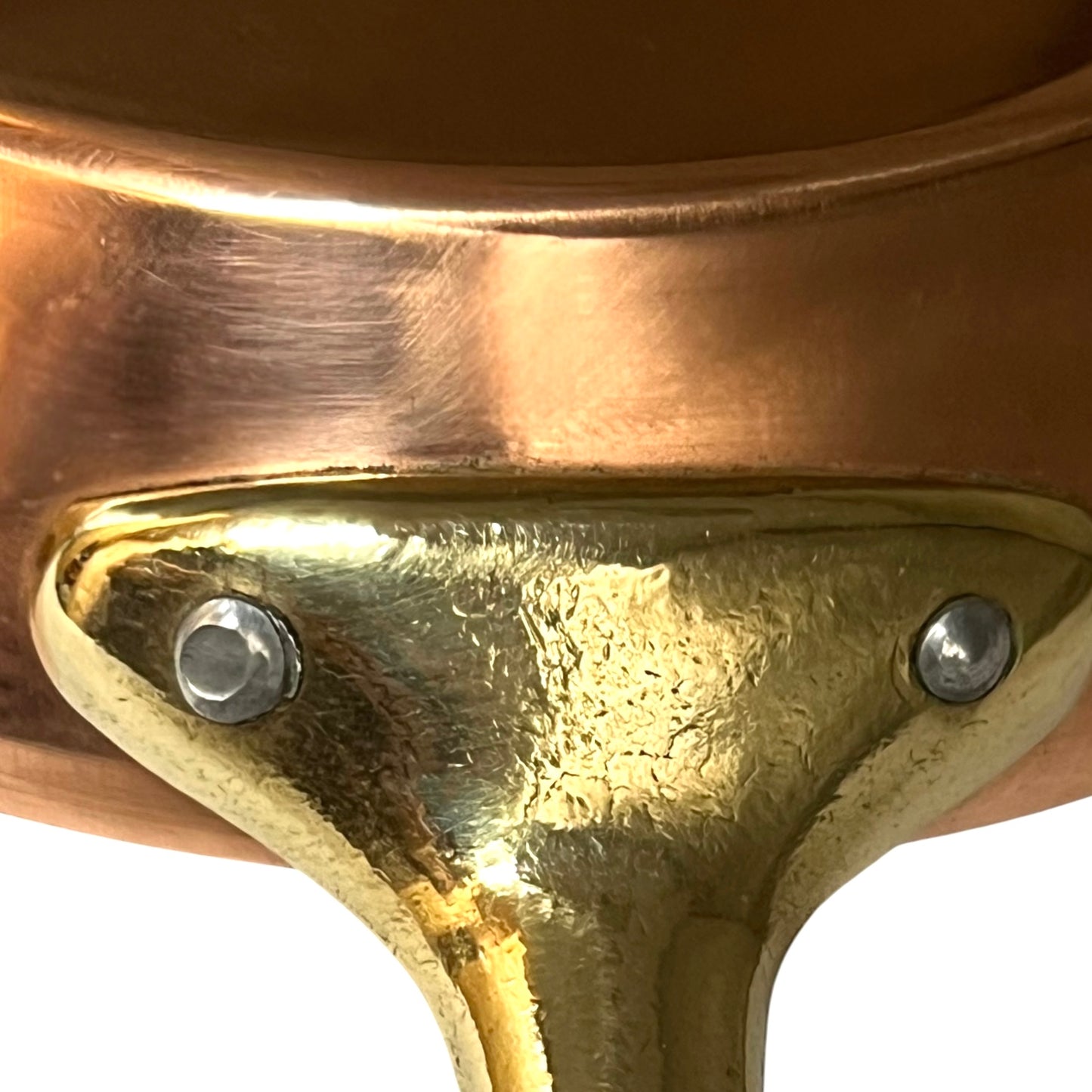 Close-up detail of a copper pot's brass or gold-toned metal rim and support bracket, secured with two silver screws, against a white background.