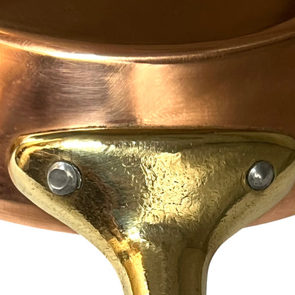 Close-up detail of a copper pot's brass or gold-toned metal rim and support bracket, secured with two silver screws, against a white background.