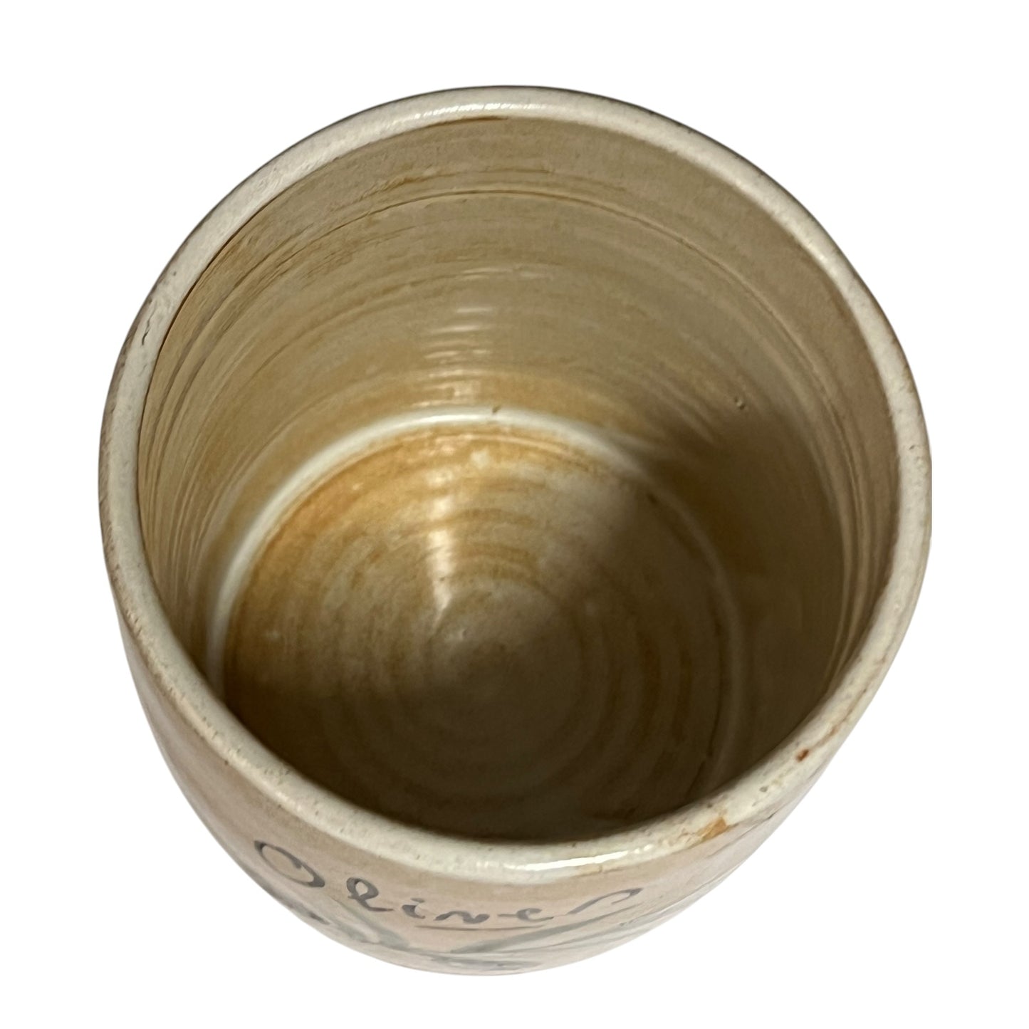A handmade ceramic cup viewed from directly above, showcasing a rustic, hand-streaked beige and brown glaze with a small dark script inscription near the bottom.