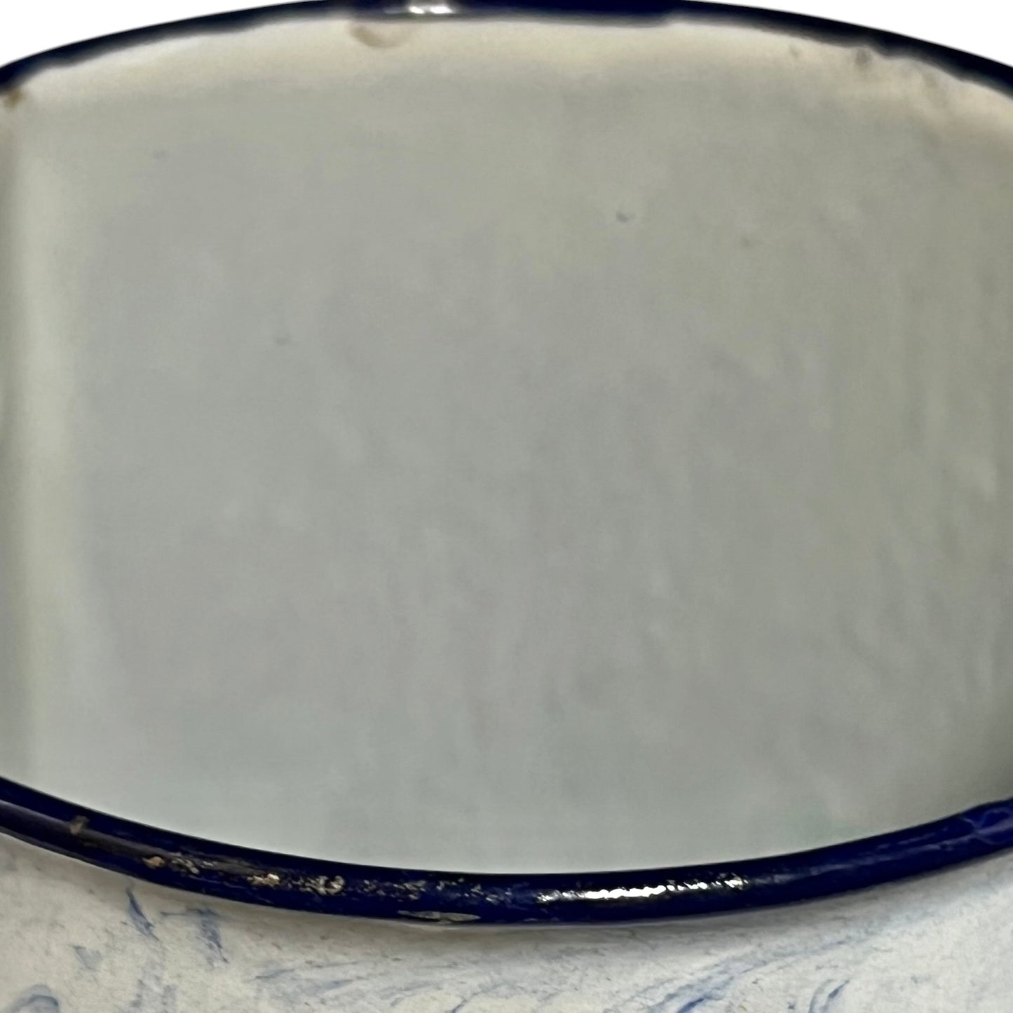 Close-up of an enamel pot with a glossy off-white interior and a dark navy blue rim, resting on a light blue marbled surface.