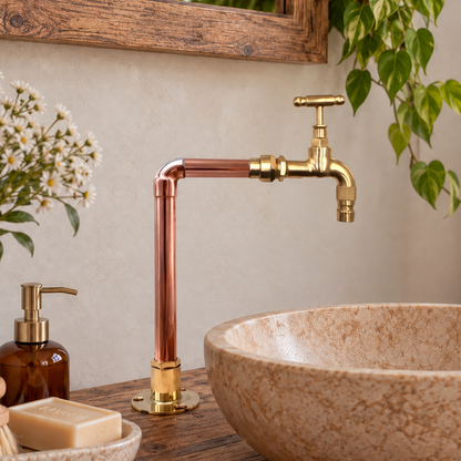 A rustic bathroom scene featuring a copper and brass gooseneck faucet mounted above a large beige stone vessel sink on a wooden vanity. The background includes a wooden mirror frame, a hanging green plant, a white pitcher of daisies, a brown soap dispenser, and a beige towel.