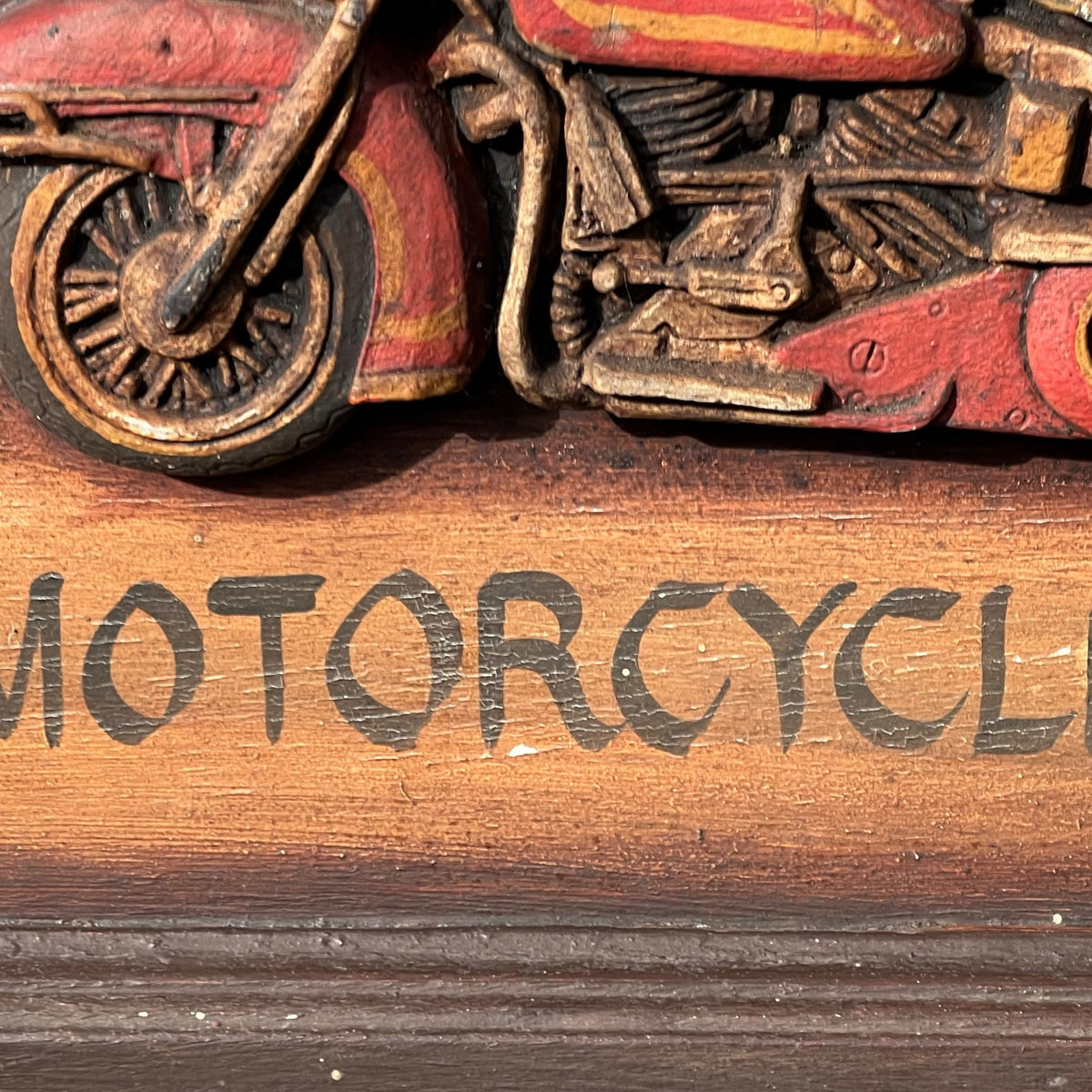 Close-up of a carved wooden motorcycle figurine with red and brown painted details, mounted above a wooden plaque engraved with the word 'MOTORCYCLE'.