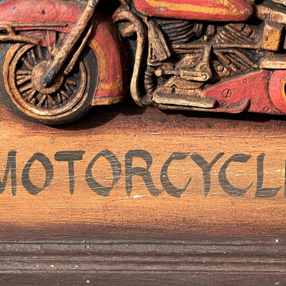 Close-up of a carved wooden motorcycle figurine with red and brown painted details, mounted above a wooden plaque engraved with the word 'MOTORCYCLE'.