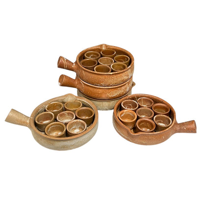 Three rustic brown ceramic cups with handles, arranged on a white background. Two cups are open, showing seven small cups inside, and one cup is closed with its handle pointing upward.