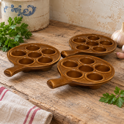 Three vintage French brown glazed ceramic escargot dishes, each featuring six individual snail compartments and a short integrated handle, arranged on a rustic wooden farmhouse table. Styled in a traditional French country kitchen setting with fresh parsley, garlic, a striped linen cloth and a blue-and-white ceramic crock. The warm caramel-brown glaze contrasts with the natural wood and neutral background. Each dish measures 14 cm in diameter and 19.5 cm including the handle.