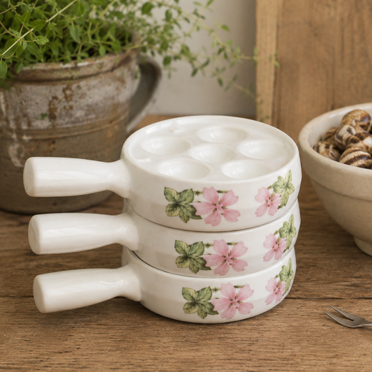 Three stacked white ceramic escargot plates with pink floral patterns and white handles, set on a rustic wooden table next to a bowl of snails.