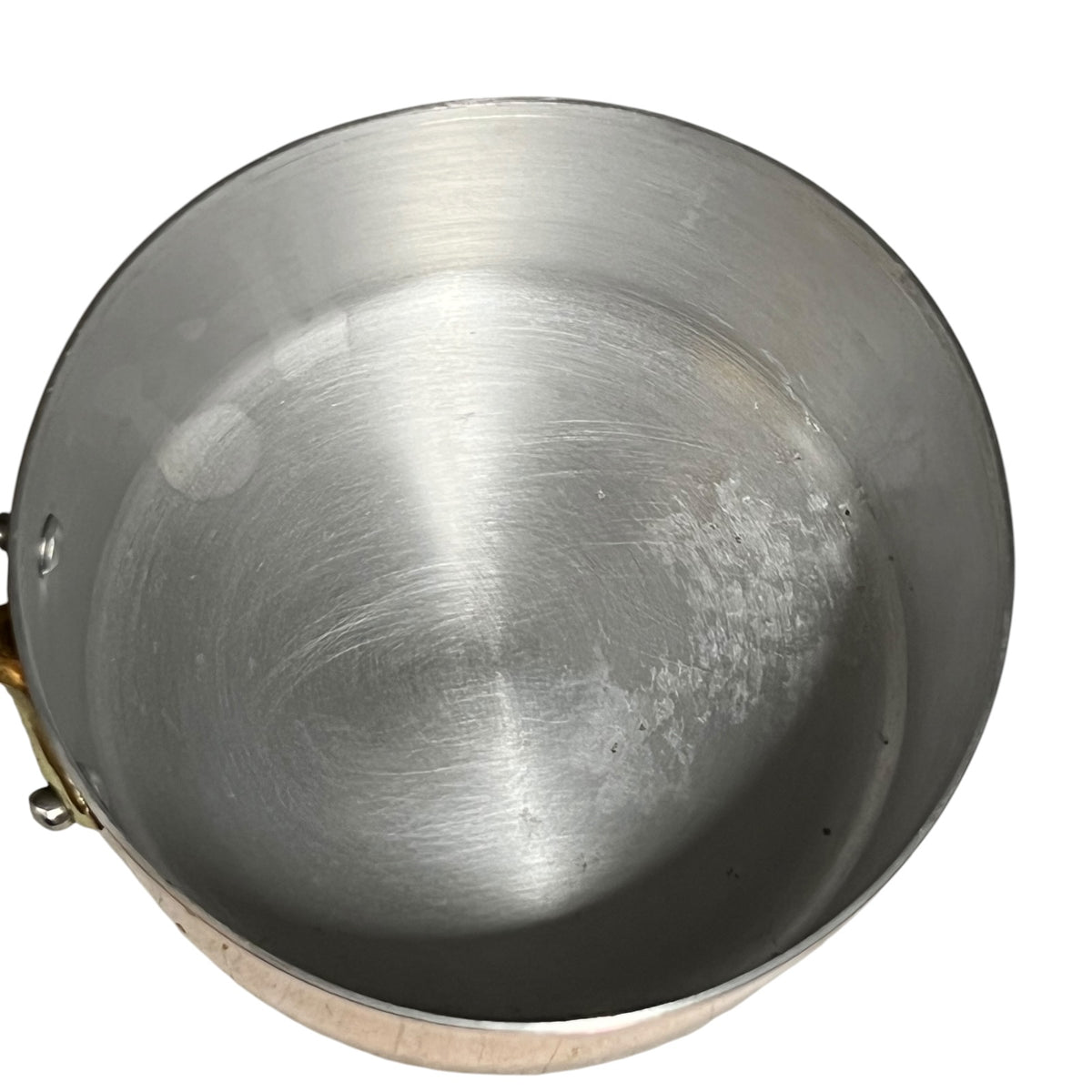 An empty aluminium lined copper saucepan viewed from directly above, showing a brushed metal interior with some light scuffs and a small brass handle attachment visible on the left