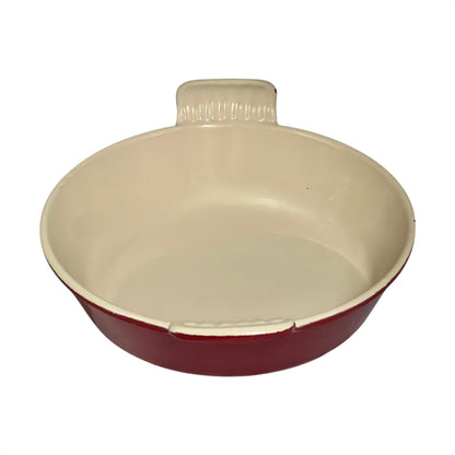 A round ceramic baking dish with a cream-colored interior and a dark red exterior, featuring a small handle on top.