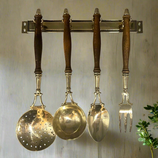Set of brass kitchen utensils with wooden handles hanging on a wall-mounted rack.