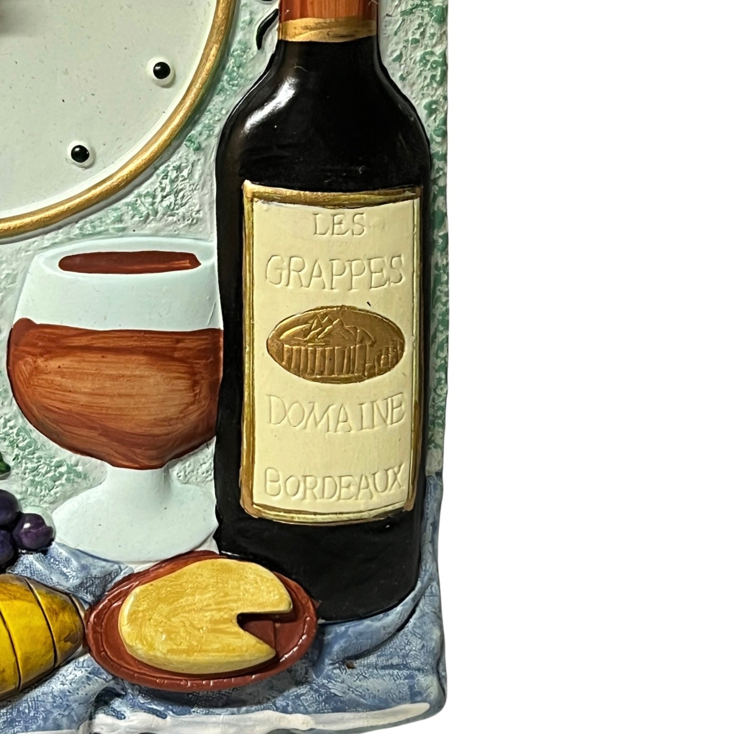 A close-up of a decorative ceramic wall plaque depicting a wine bottle labeled 'Les Grappes Domaine Bordeaux,' a wine glass, grapes, cheese, and a loaf of bread on a textured background.