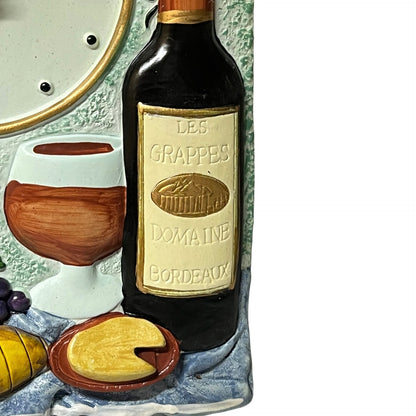 A close-up of a decorative ceramic wall plaque depicting a wine bottle labeled 'Les Grappes Domaine Bordeaux,' a wine glass, grapes, cheese, and a loaf of bread on a textured background.