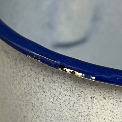 Close-up of an enamel pot, showing a blue rim with chipped enamel revealing silver metal underneath, set against a speckled off-white ceramic body.