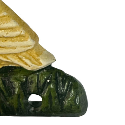 Close-up of a small decorative figurine or ornament showing a beige textured roof-like section resting on a dark green base with a hole, photographed against a plain white background.