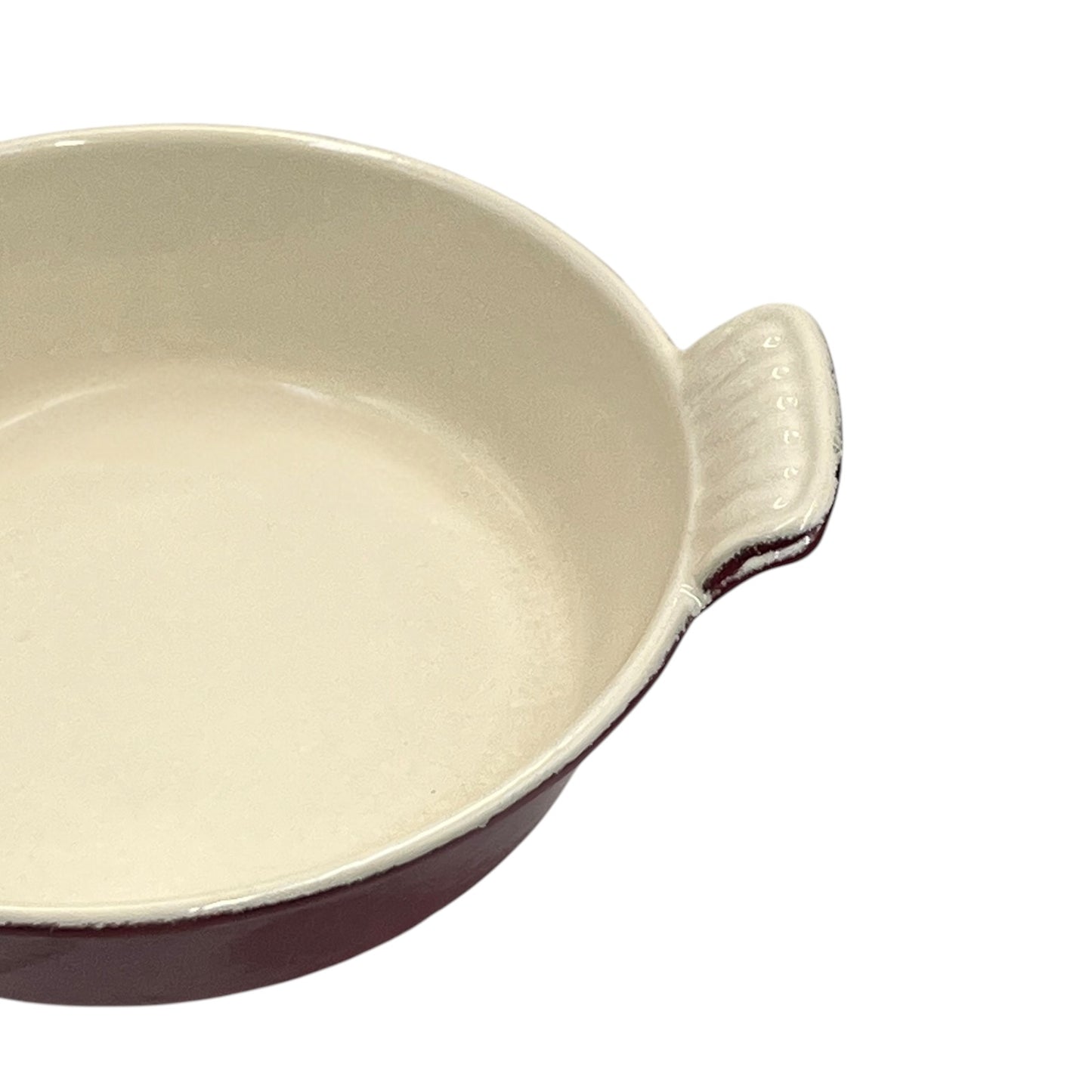 A close-up of a round ceramic baking dish with a cream-colored glazed interior and a dark reddish-brown exterior, featuring a small ribbed handle on the right side, set against a plain white background.