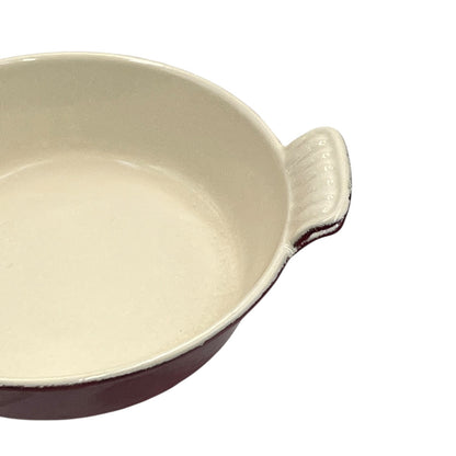 A close-up of a round ceramic baking dish with a cream-colored glazed interior and a dark reddish-brown exterior, featuring a small ribbed handle on the right side, set against a plain white background.