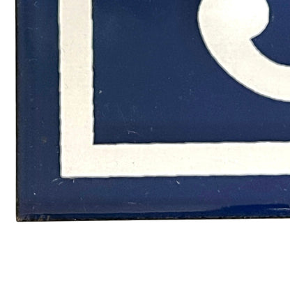 Close-up of a dark blue decorative tile or sign with a white border and partial white graphic, shown against a white background.