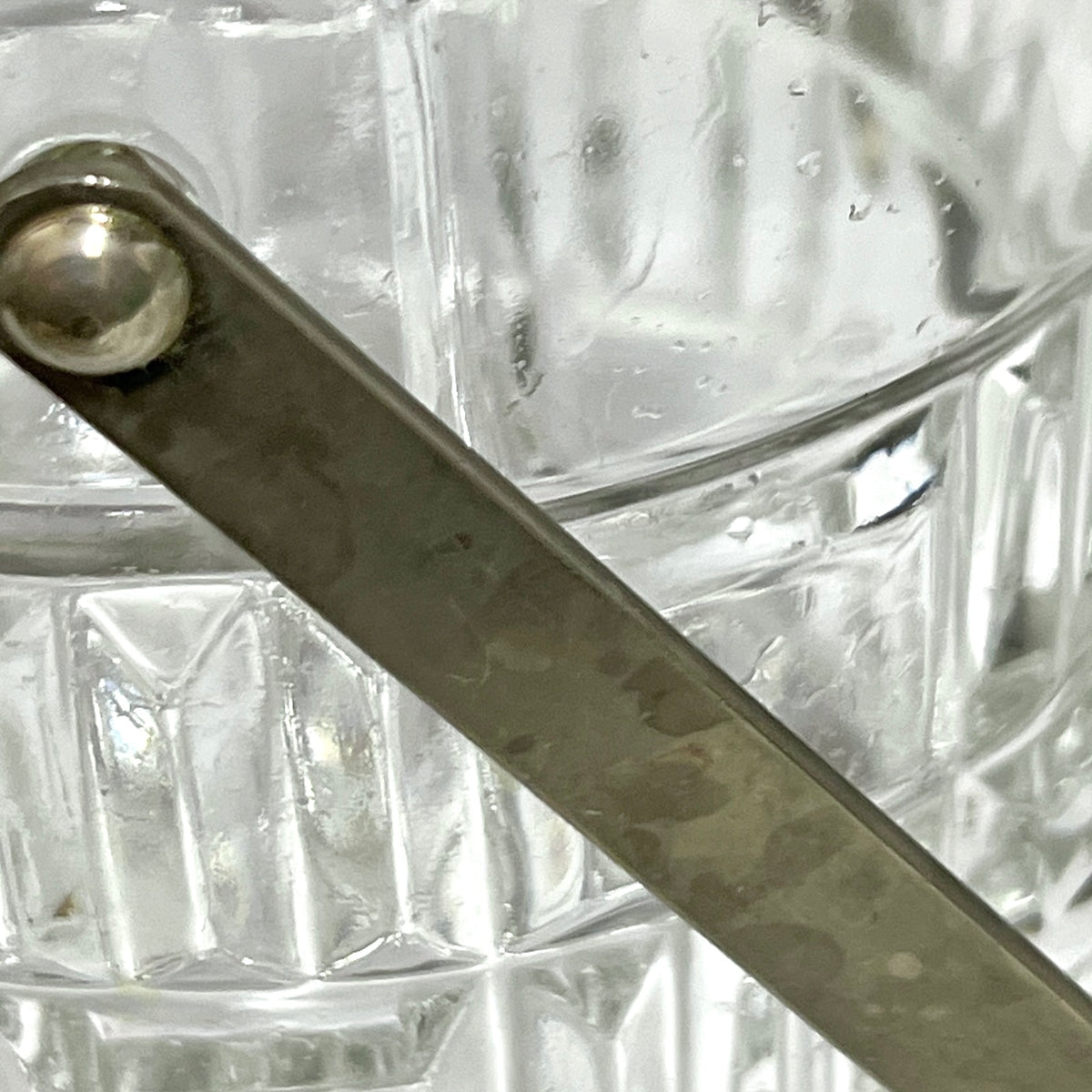 Close-up of a clear textured glass container with a hammered silver metal handle attached near the rim.