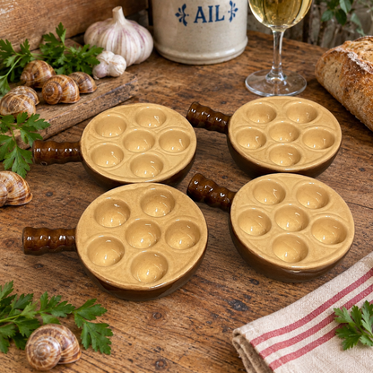 Four round ceramic escargot dishes with handles, arranged on a rustic wooden table with garlic, snails, parsley, bread, and a glass of white wine in the background.