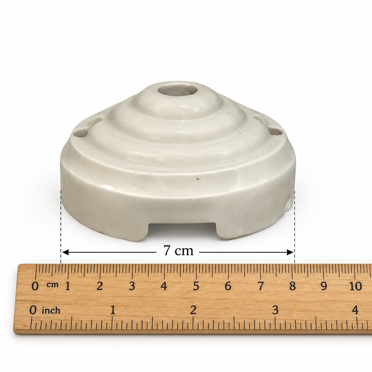 A small off-white ceramic or porcelain ceiling rose with a rounded, tiered top and side holes, shown above a wooden ruler with a 7 cm dimension marker on a white background.