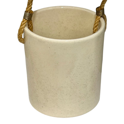 A cylindrical ceramic utensil pot in a speckled cream-beige glaze, featuring a natural jute rope hanging loop with brass wire wraps on the sides, shown against a plain white background.