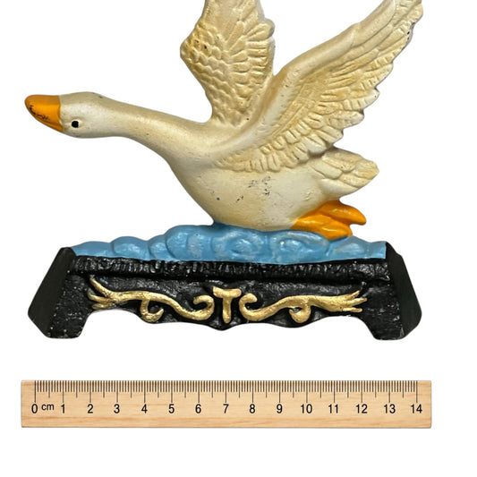 A decorative goose doorstop with wings raised, shown on a black base with gold scrollwork and blue water details. A wooden ruler is placed below the product to indicate scale.