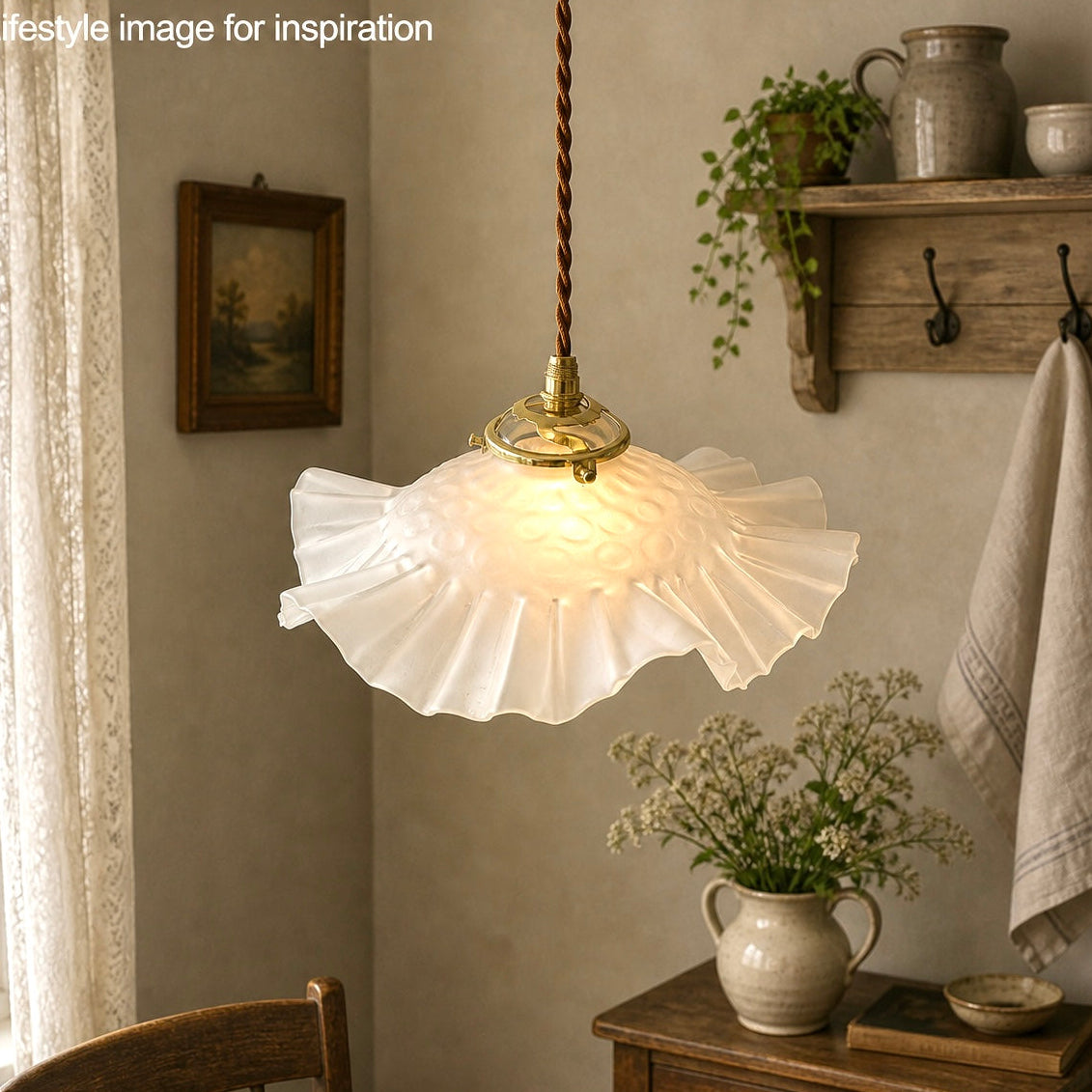 French vintage frosted glass lampshade with a frilled scalloped style edge and a brass collar suspended with vintage wiring. In the background there is a wooden table and wall hooks holding a towel, plants and an open window with the top of a chair just visible giving a farmhouse kitchen style