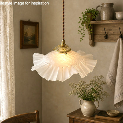 French vintage frosted glass lampshade with a frilled scalloped style edge and a brass collar suspended with vintage wiring. In the background there is a wooden table and wall hooks holding a towel, plants and an open window with the top of a chair just visible giving a farmhouse kitchen style