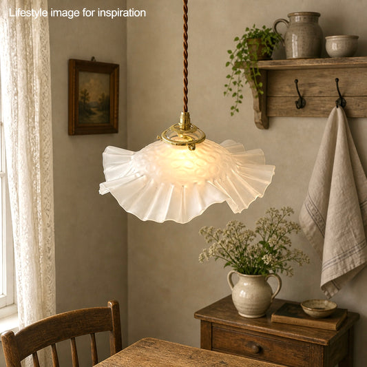 French vintage frosted glass lampshade with a frilled scalloped style edge and a brass collar suspended with vintage wiring. In the background there is a wooden table and wall hooks holding a towel, plants and an open window with the top of a chair just visible giving a farmhouse kitchen style