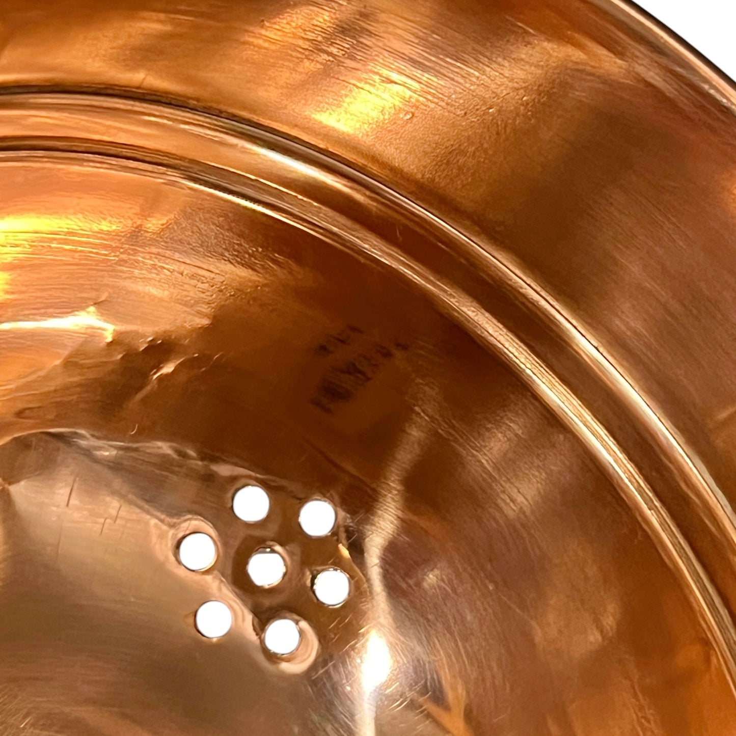 Close-up of the inside of a shiny copper-colored metal bowl or pot, showing a perforated drain hole and warm reflective highlights.