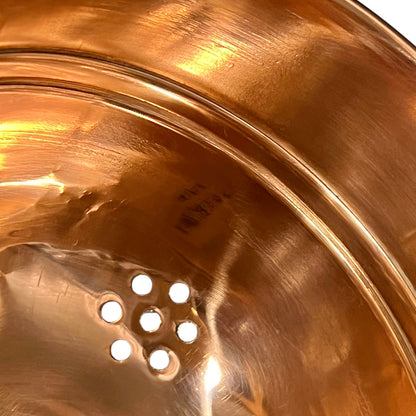 Close-up of the inside of a shiny copper-colored metal bowl or pot, showing a perforated drain hole and warm reflective highlights.