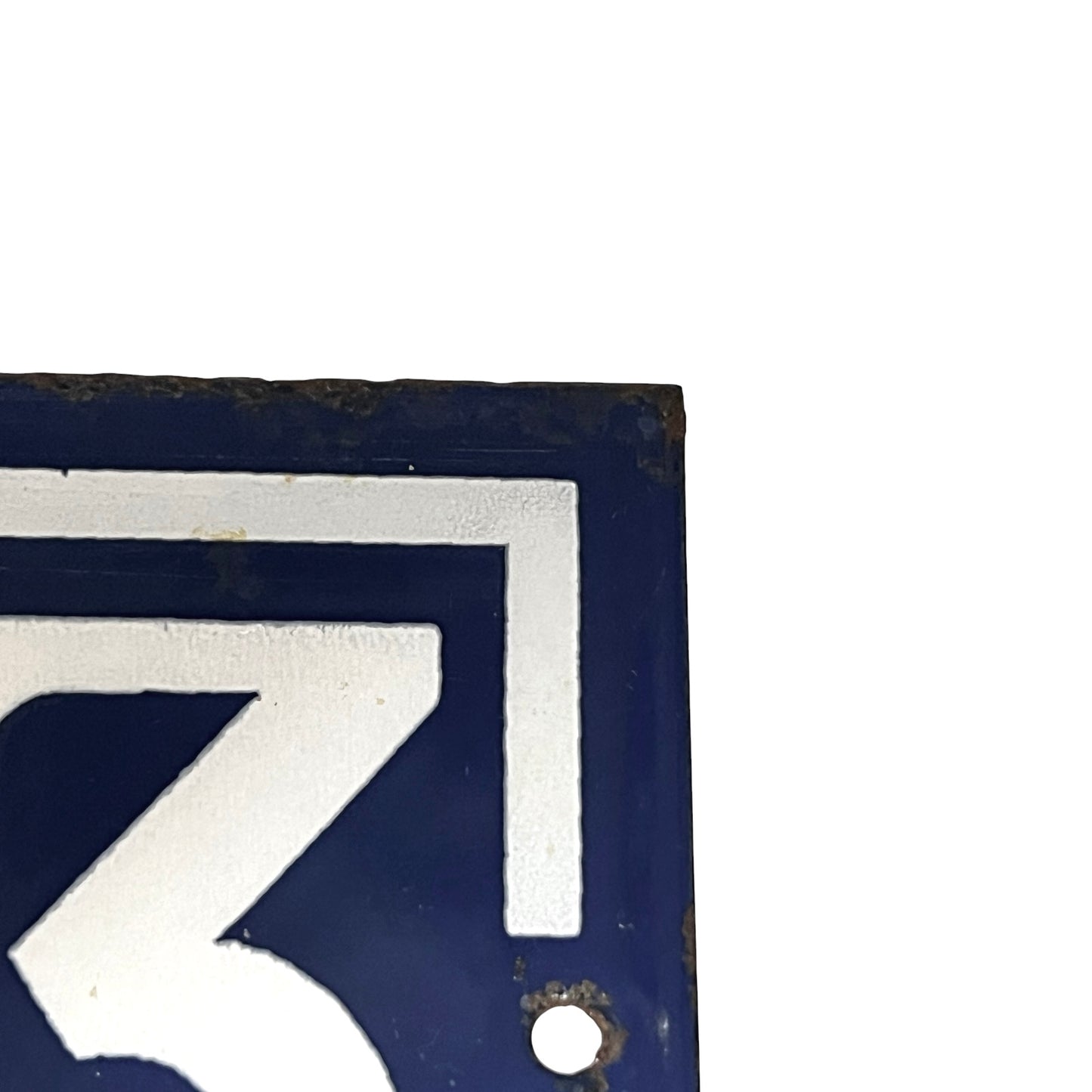 A close-up corner of a dark blue enamel-style sign with a white geometric numeral or letterform and a small round hole near the bottom, shown against a white background.
