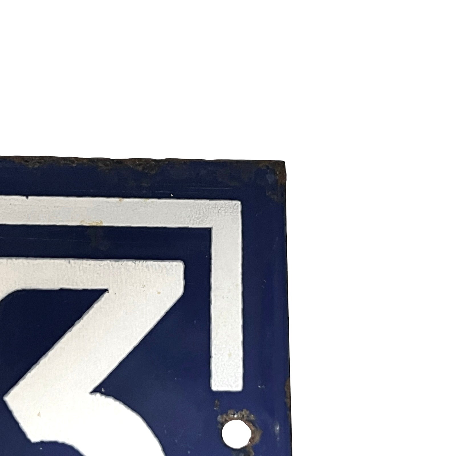 A close-up corner of a dark blue enamel-style sign with a white geometric numeral or letterform and a small round hole near the bottom, shown against a white background.