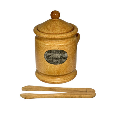 A small ceramic canister with a lid and wooden knob, shown on a white background with a wooden spoon or spatula placed below it. The canister has a speckled tan-brown glaze and a round label reading 'Cranberries'.