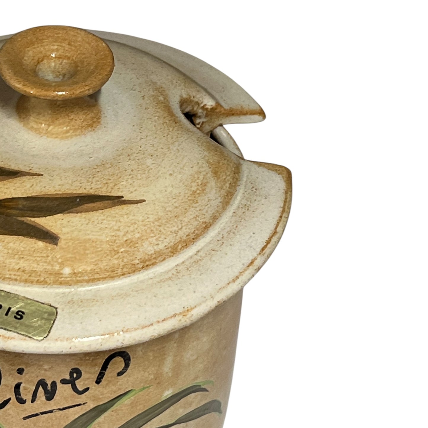 Close-up of a ceramic lidded container or jar with a rounded knob, beige and cream glaze, hand-painted green leaf details, and partial handwritten text on a white background.