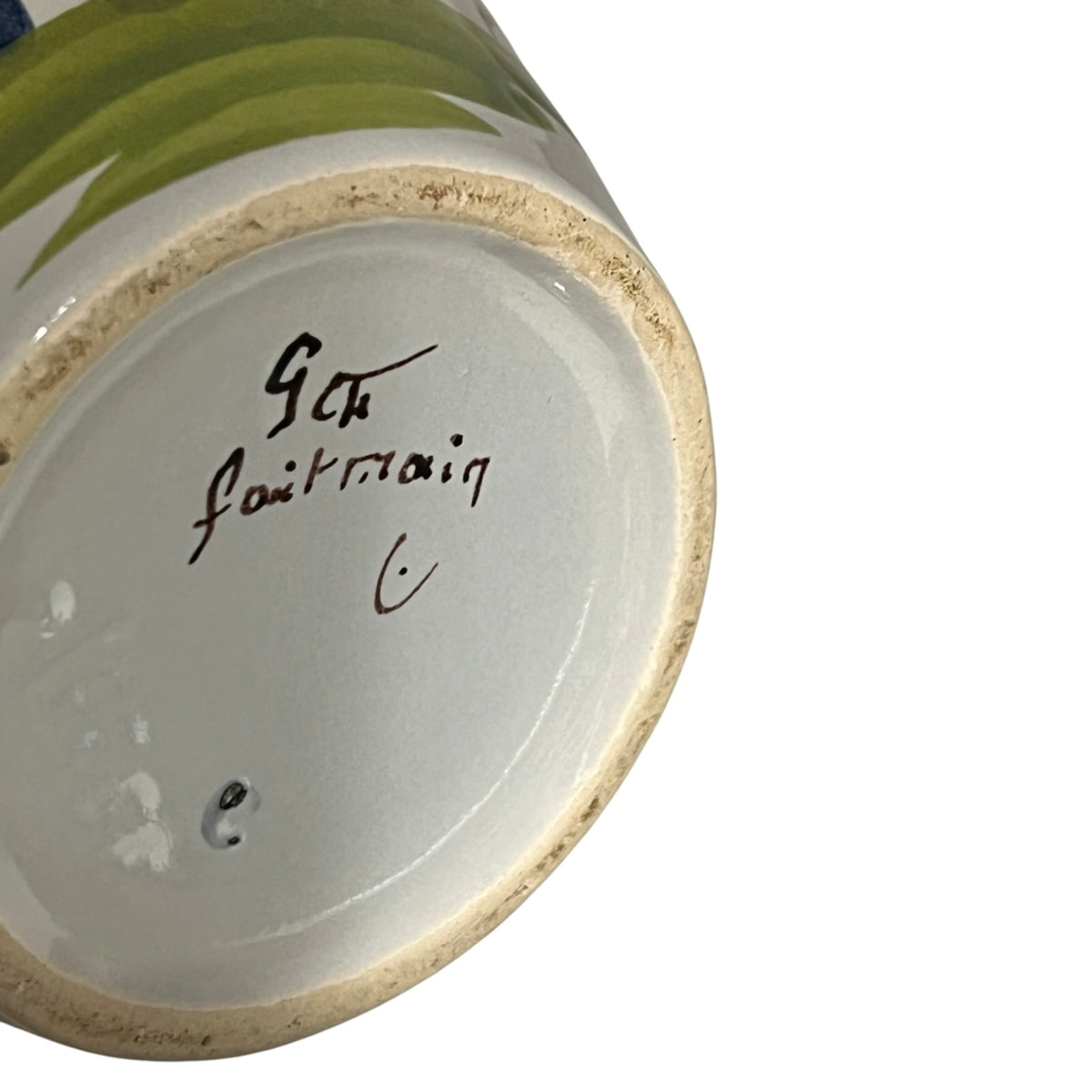 Close-up of the bottom of a ceramic mug or vase showing a white glazed interior with a handwritten signature and a natural beige clay rim. A partial green painted motif is visible along the upper edge against a white background.