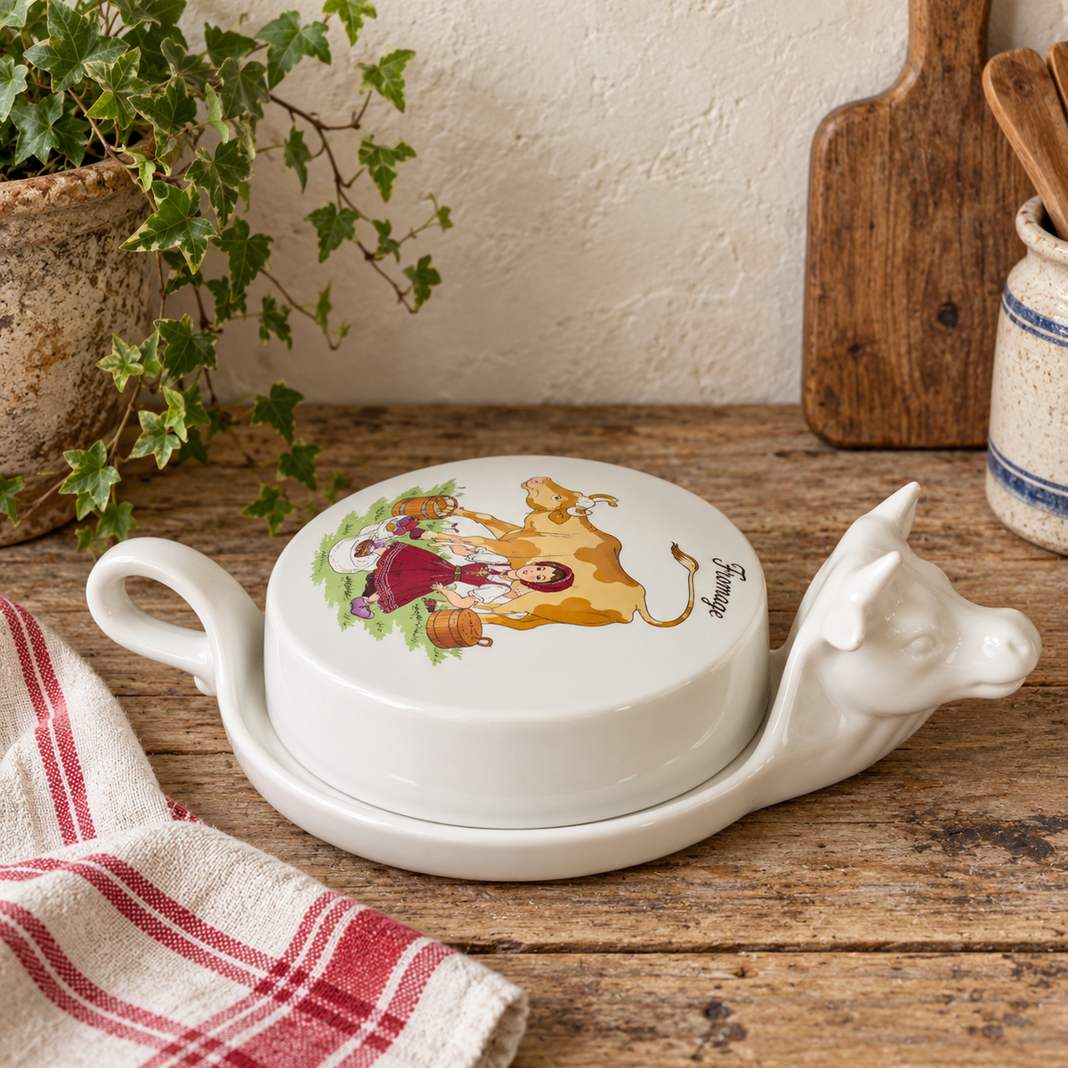 Vintage French L'Hirondelle porcelain cheese dish with cow-shaped white base and illustrated Fromage lid, traditional French farmhouse cheese server shown on a wooden top beside a ed and white tea towel